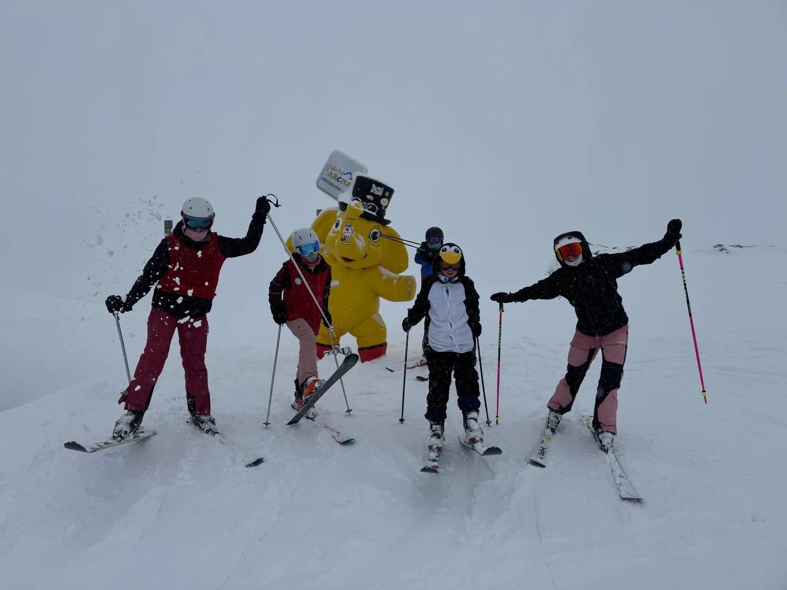 Vijf skiërs op de piste met de armen omhoog, een van hen in een geel pak