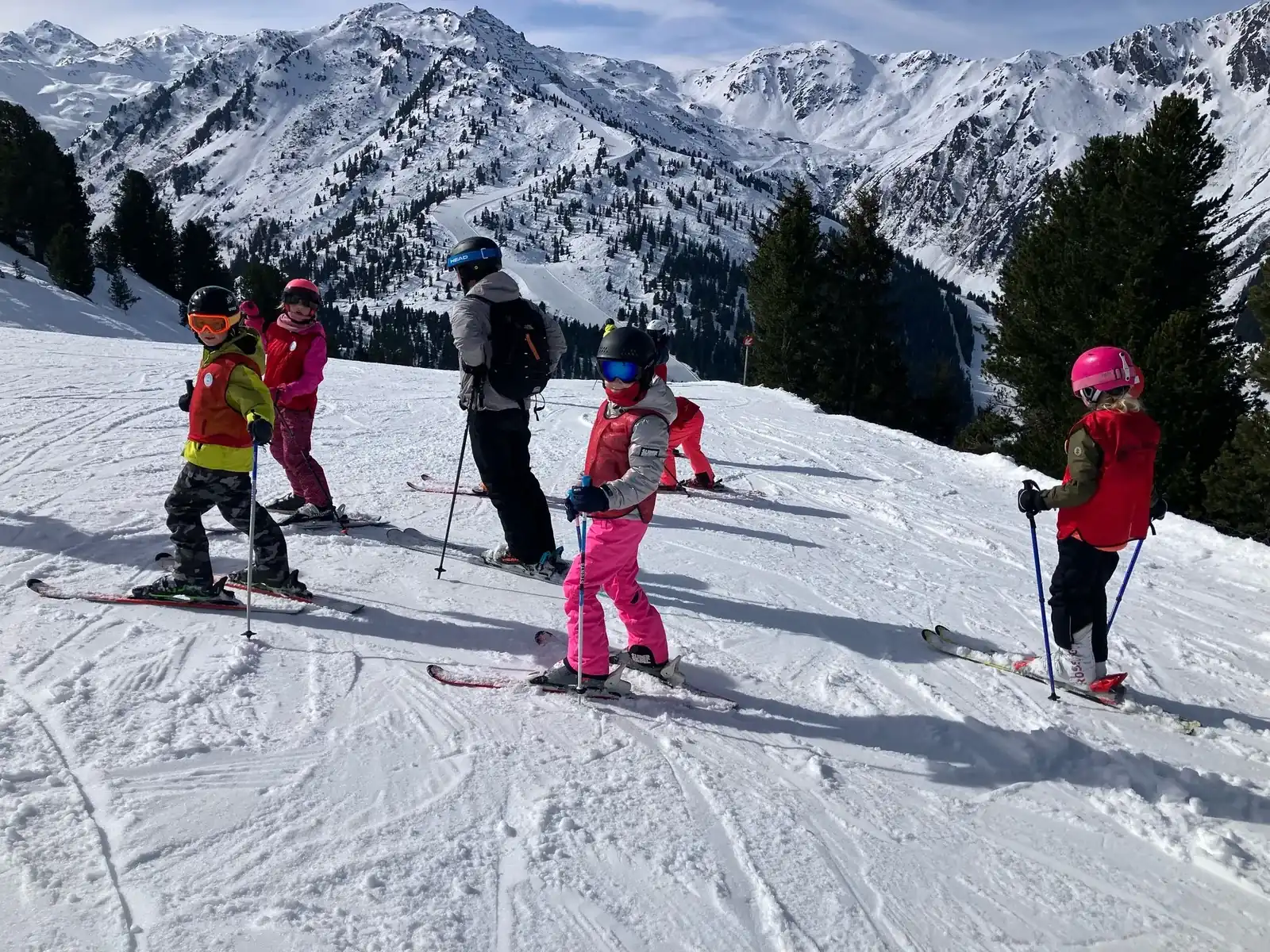 Kinderen oefenen op een brede piste, met een skileraar ernaast en bergen erachter